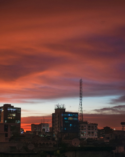 A view of the modern Dhaka skyline at sunset, with tall buildings silhouetted against an orange sky.
